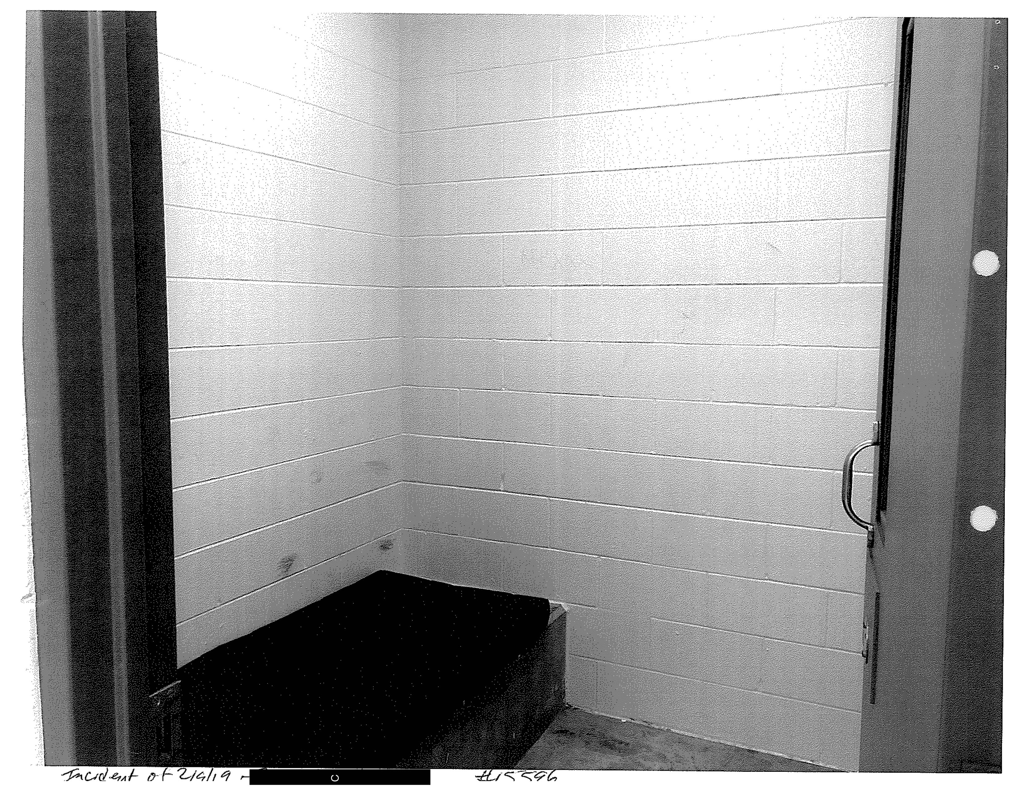 Black and white photo of a small cell with light walls, a dark bed on the left side and a door that opens inward.