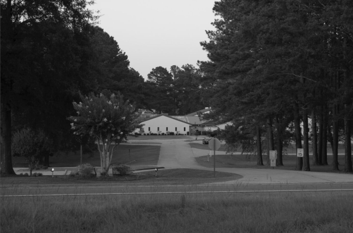 Black and white photo of a low building complex in the background, surrounded by forest. A road leads up to the building.