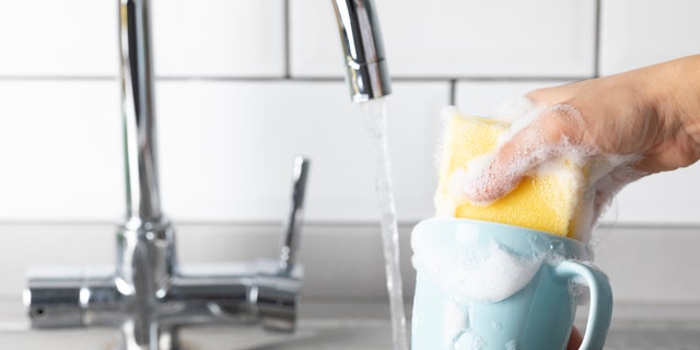 This file image shows a woman washing a coffee cup in the kitchen sink. 