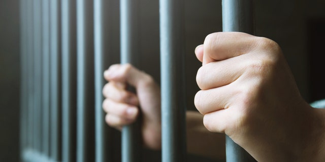 A man in prison puts his hands around the steel bars of a jail cell.