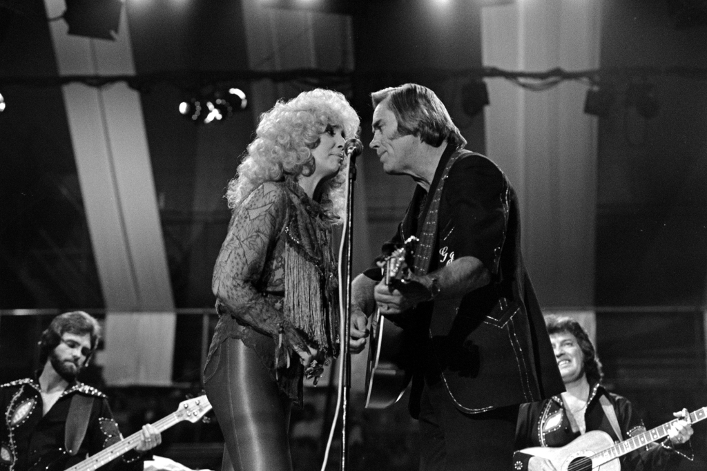 Tammy Wynette and George Jones performing together in London in 1981. Tammy has big blond curls and is wearing leather pants; George is dressed in black and is strumming an acoustic guitar. They're both very close to each other while singing into the microphone.