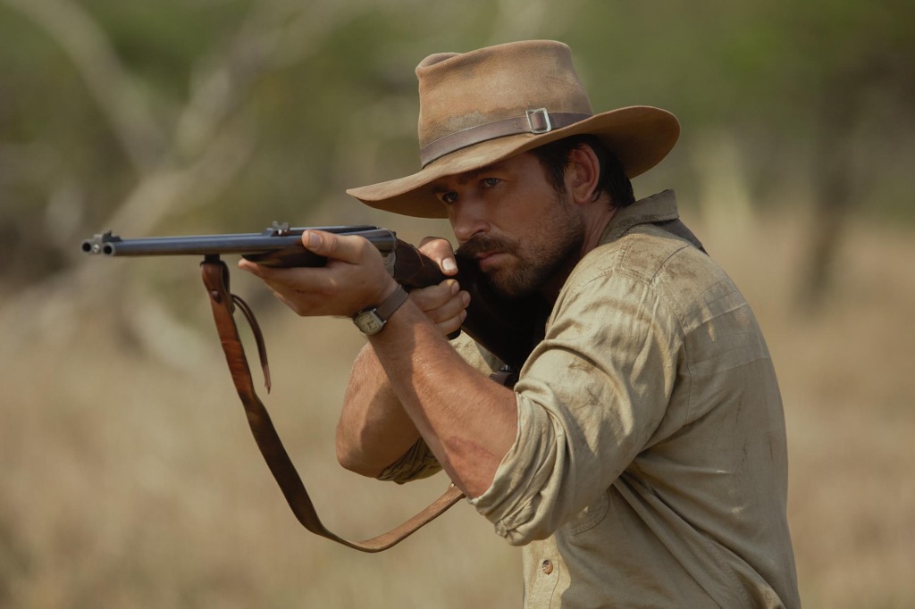 Brandon Sklenar as Spencer Dutton. He's in the African bush and is pointing a rifle and ready to shoot. He's wearing a brown safari hat and khaki shirt.