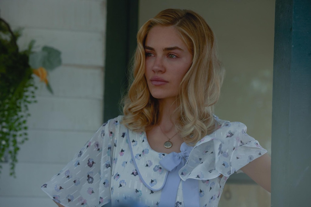 Michelle Randolph as Elizabeth Stafford. She's on a porch and is wearing a white dress with a blue sash and blue flowers on the dress. She's got blonde hair.