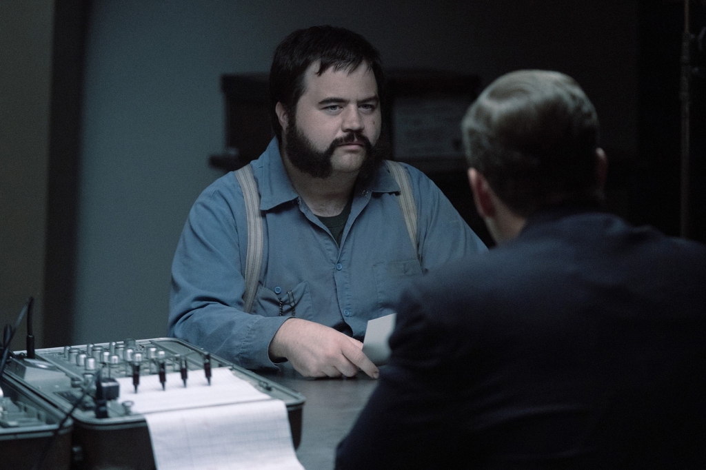Paul Walter Hauser as serial killer Larry Hall in "Black Bird." He's got long muttonchop sideburns and is wearing a blue shirt with grey suspenders while taking a lie-detector test.