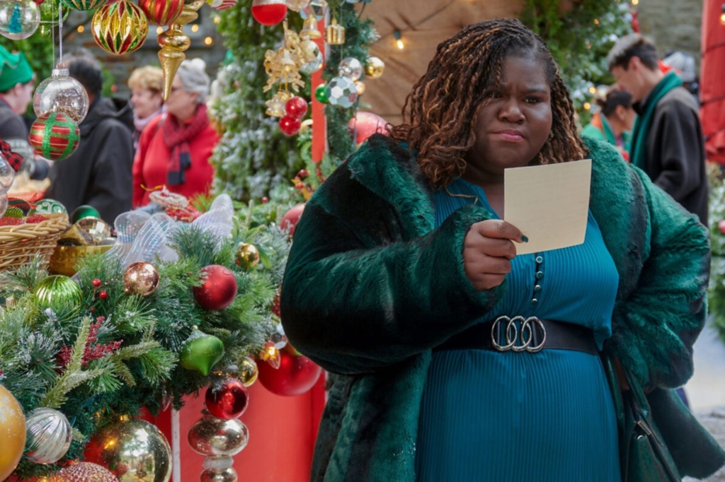 Gabourey Sidibe in "All I Didn't Want For Christmas." She's standing in front of a Christmas tree and looking at a note she's holding in her hand and frowning.