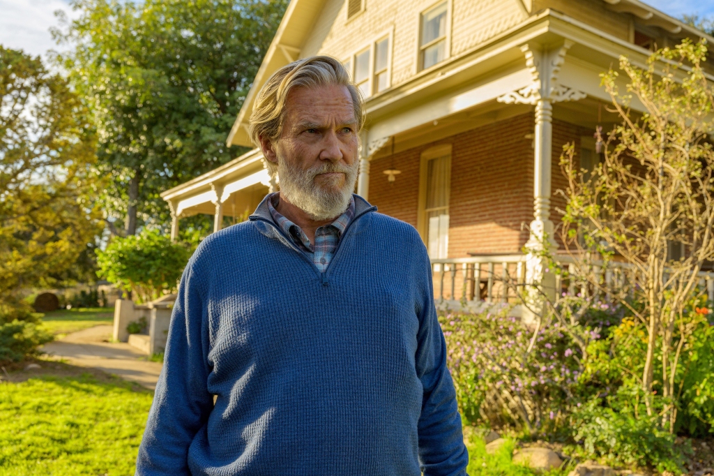 Jeff Bridges as Dan Chase in "The Old Man." He's standing outside of his house in the daylight and looking off-camera. He's wearing a blue sweater. He has a grey beard.
