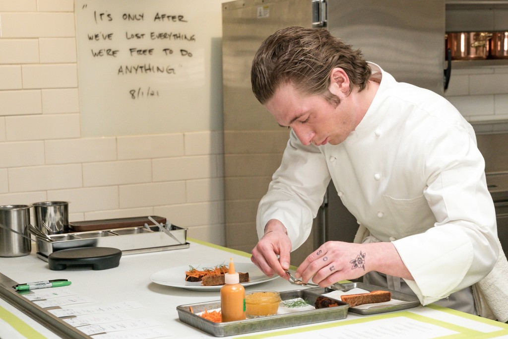Jeremy Allen White in "The Bear." He's in the kitchen wearing a white chef's uniform as he looks down and carefully prepares a dish with an intense look on his face. He's got tattoos on his left fingers and wrist." 