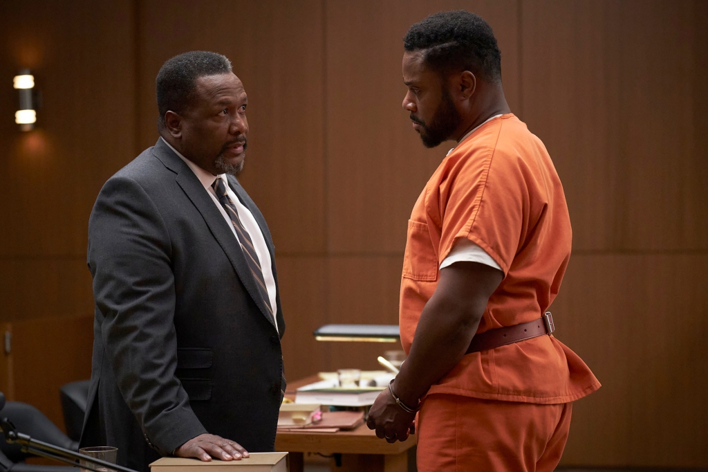 Wendell Pierce wearing a suit standing across a table from Malcolm-Jamal Warner, who is in an orange prison jumpsuit. 