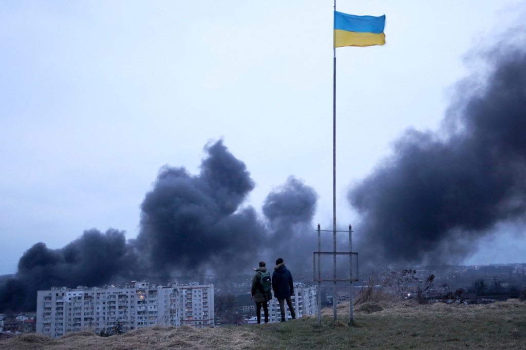 People standing near a Ukrainian national flag watch as dark smoke billows following an air strike in the western Ukrainian city of Lviv.