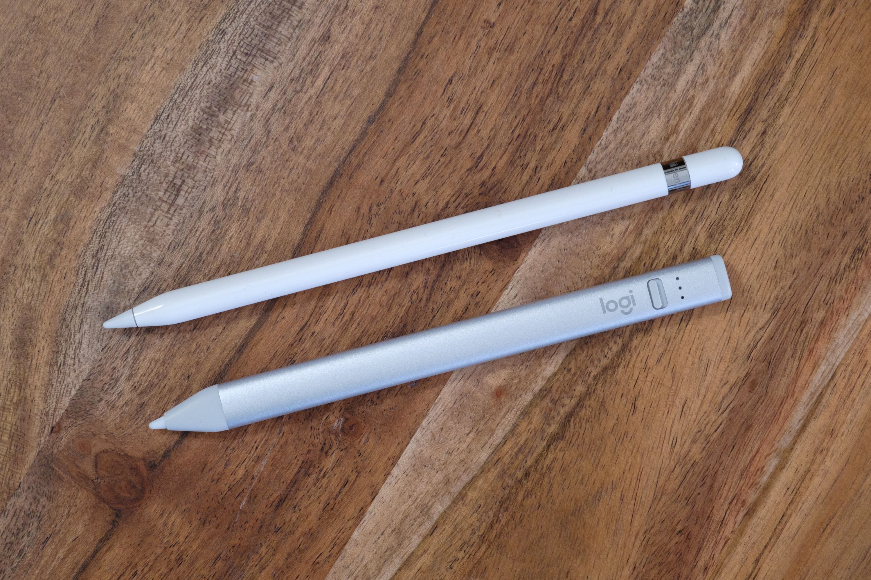 A Logitech Crayon next to a 1st Gen Apple Pencil on a wood table, viewed from the top down.