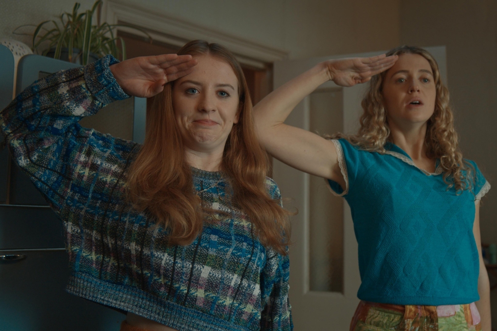 Carrie (Sofia Oxenham) right, and Jen (Mairead Tyers, left, in "Extraordinary" stand next to each other smiling and saluting. 