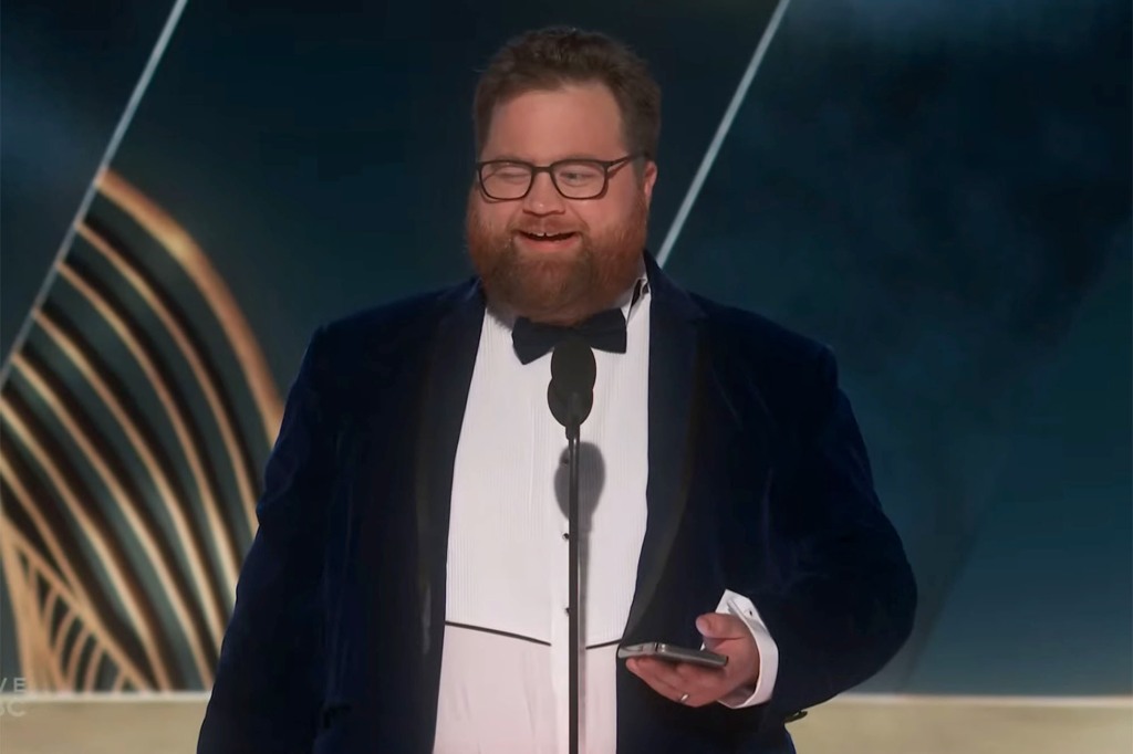 Paul Walter Hauser on stage. He's wearing a tuxedo and holding his cell phone. He's smiling and is wearing glasses and has a beard.
