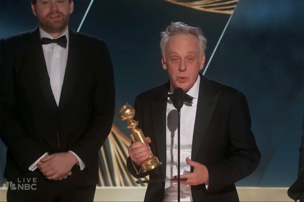 Mike White on stage, holding his Golden Globe Award. He's wearing a tuxedo and has gray hair.