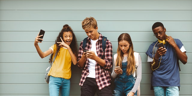 College friends standing outdoors looking using their cell phones. Young girl taking a selfie standing outdoors against a wall with her friends looking at their mobile phones. (iStock)