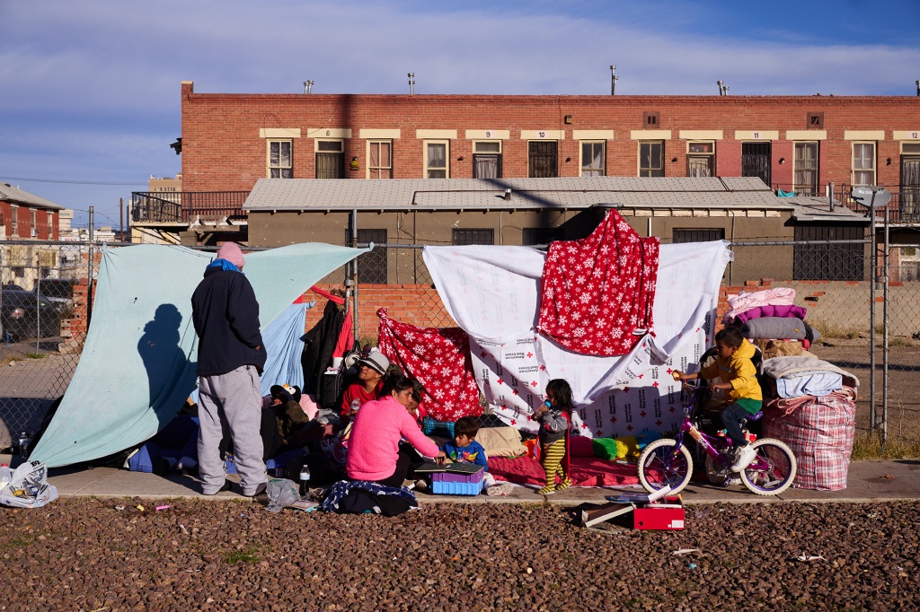 sheets are hung from the wire fence to offer shelter for a family, a mother is showing a child a book, while another rides a bike in the background. The father talks to a man in a dark hoodie on the sidewalk  