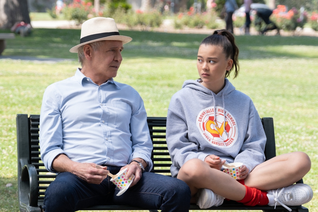 Harrison Ford and Lukita Maxwell as Paul and Alice in "Shrinking." They're sitting side-by-side on a park bench in the bright sunshine. Paul is eating ice cream and wearing a white Panama hat; Alice is also eating ice cream and is wearing a sweatshirt with her high school's name across the front.