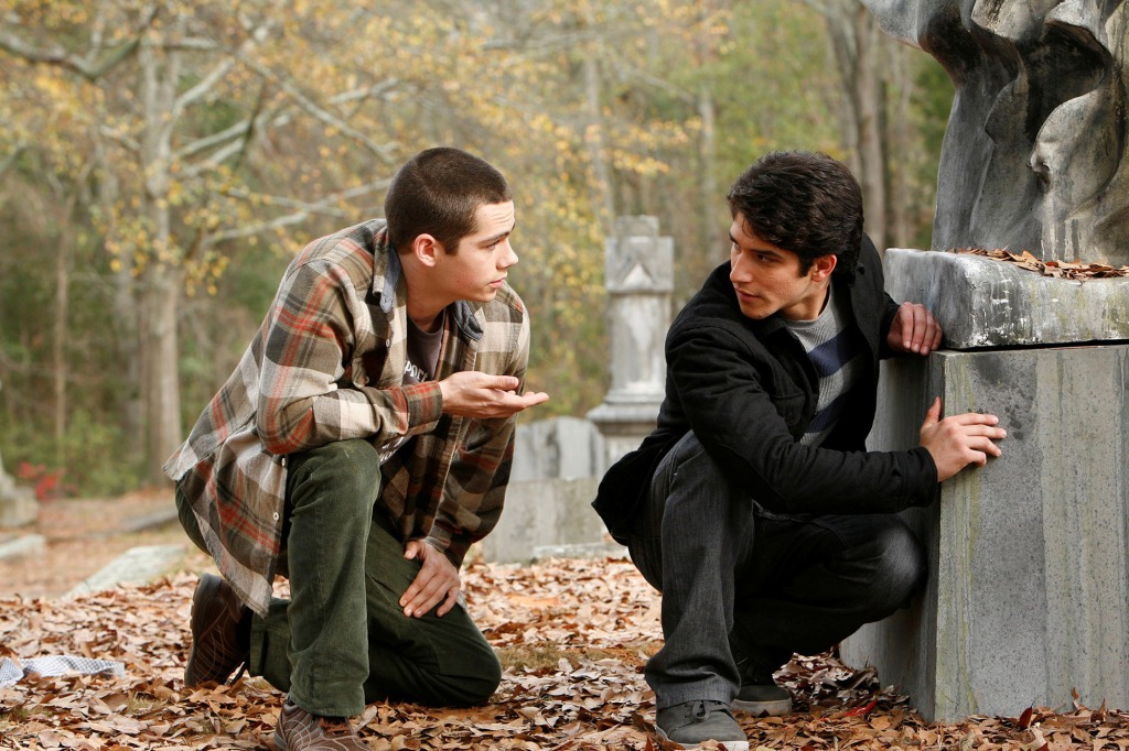 Dylan O'Brien and Tyler Posey in "Teen Wolf" crouching next to a grave looking at each other. 