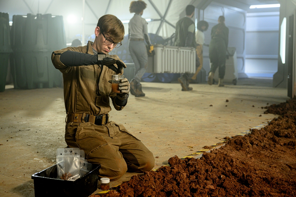 Ryan Adams as Angus Medford in a scene from "The Ark." He's kneeling in front of a pile of topsoil and is putting some of that into a glass jar. He's wearing a green jumpsuit.