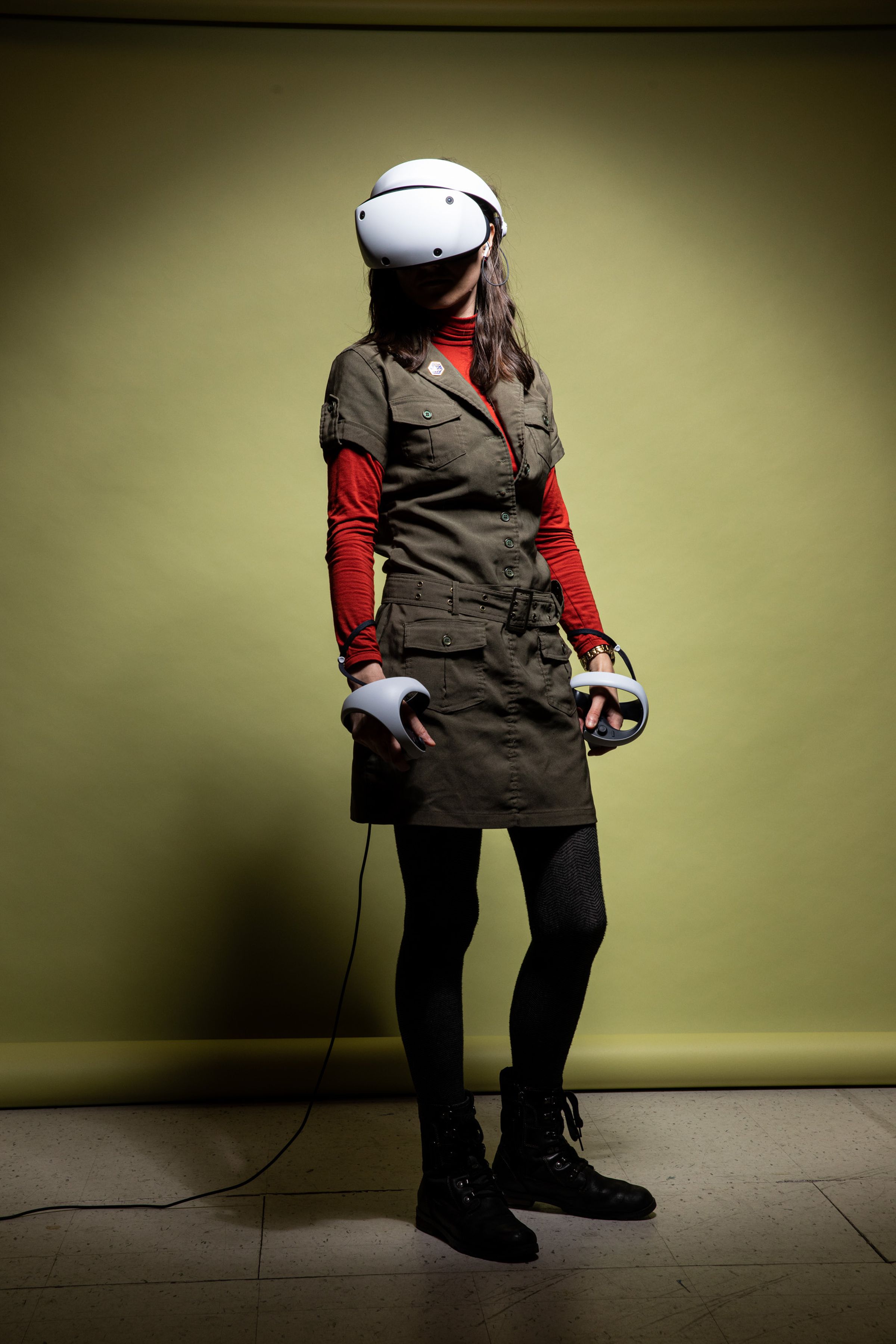 A woman stands wearing the PSVR 2 headset and controllers, with a cord dangling down and across the floor.