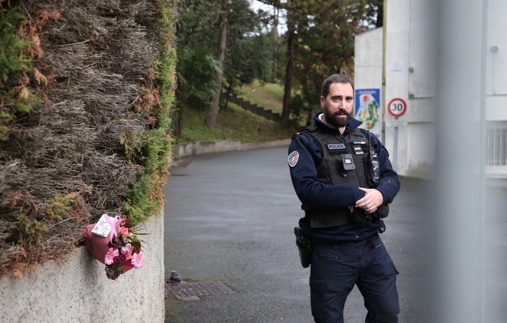 A police officer guards the entrance to a private Catholic school next to a bouquet of flowers.