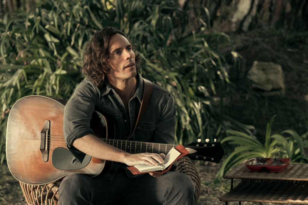 Sam Claflin sitting in a garden with a guitar. 