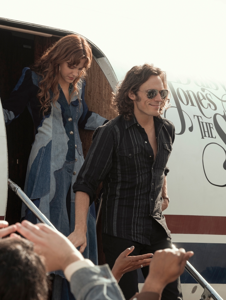 Riley Keough and Sam Claflin smiling exiting a plane. 