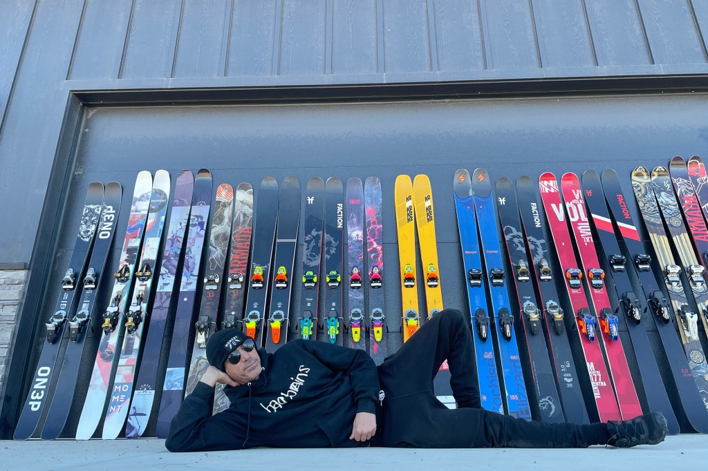 Man wearing black winter clothing lying down propped up on elbow in front of skis lined up.