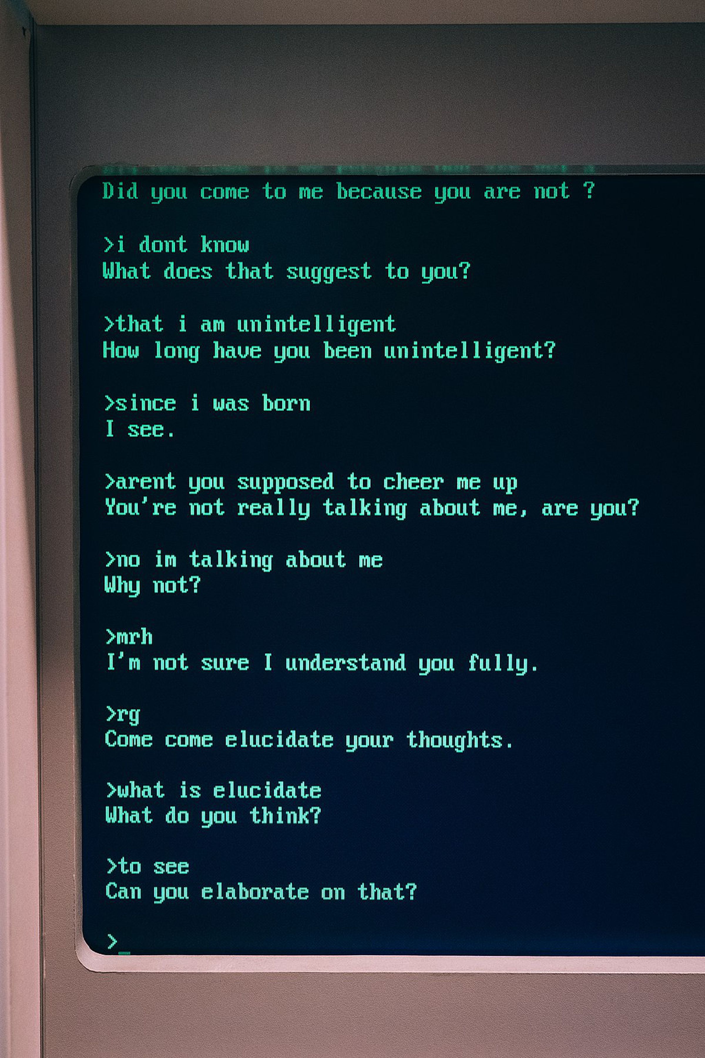 A photograph of an old fashioned computer monitor showing a conversation with ELIZA. The chatbot asks questions like “Can you elaborate on that?”