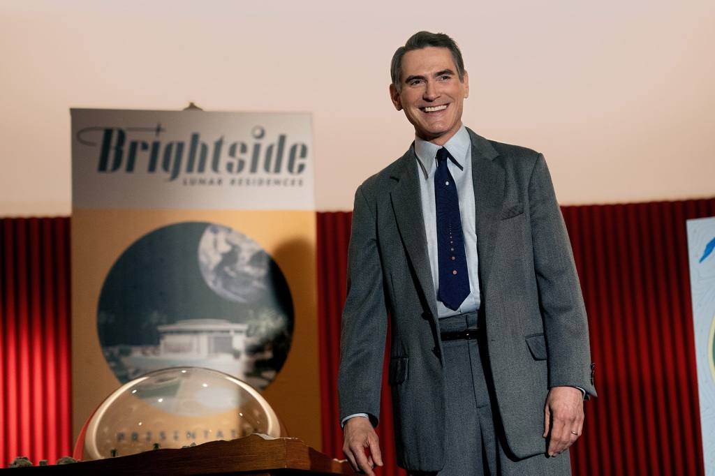 Billy Crudup in "Hello Tomorrow!" wearing a suit and smiling standing in front of a poster board. 