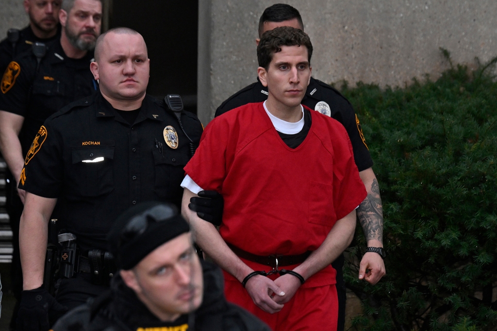 Bryan Kohberger wearing a red prison jumpsuit is escorted by officers from court into a waiting police vehicle. His handles are shackled.