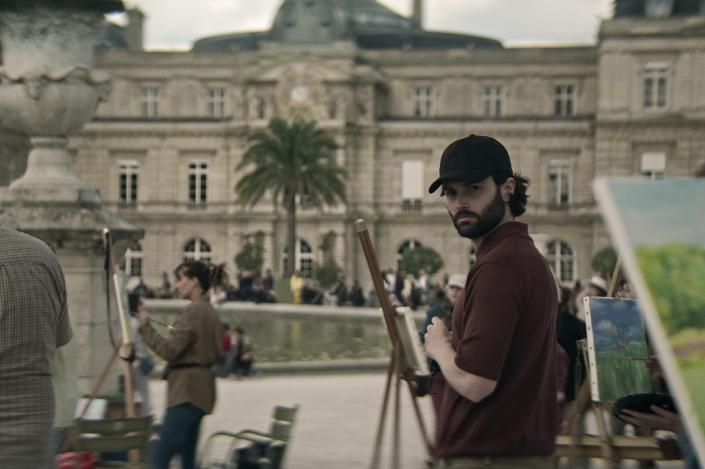 Penn Badgley as Joe Goldberg in "You" wearing a baseball hat painting in front of a palace. 