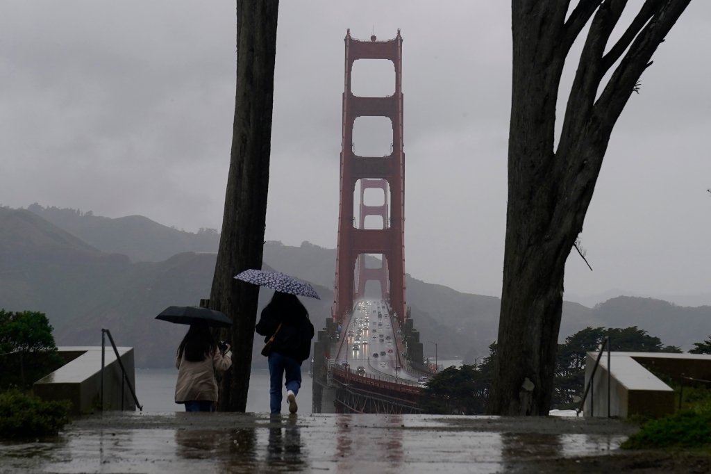 Traffic moves on the Golden Gate Bridge as people carry umbrellas while walking down a path at the Golden Gate Overlook in San Francisco, Calif., on March 9, 2023.