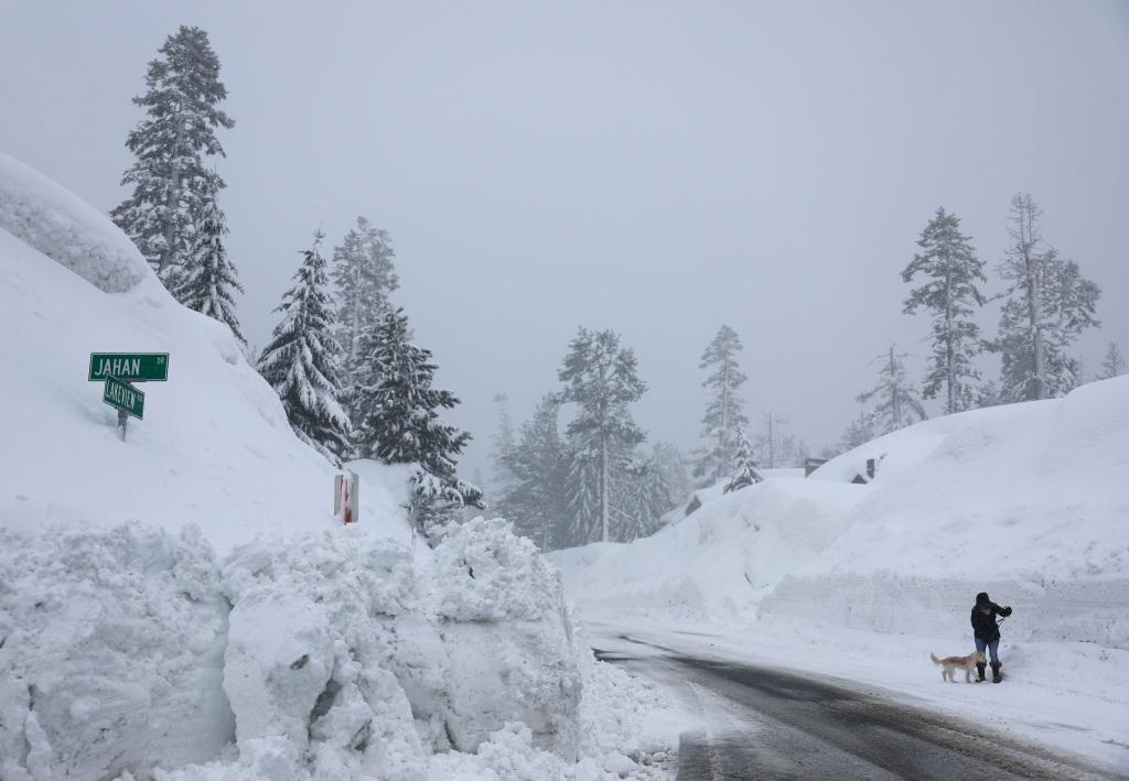 A person walks a dog near snowbanks piled up from current and previous storms as snow continues to fall in the Sierra Nevada mountains.