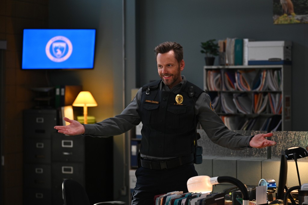 Joel McHale smirking and holding his arms out standing in an office. 