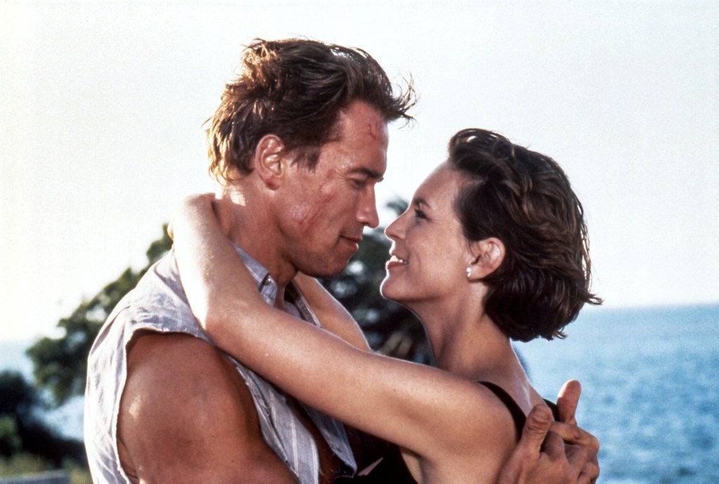 Arnold Schwarzenegger and Jamie Lee Curtis in "True Lies" embracing and smiling at each other by the water. 