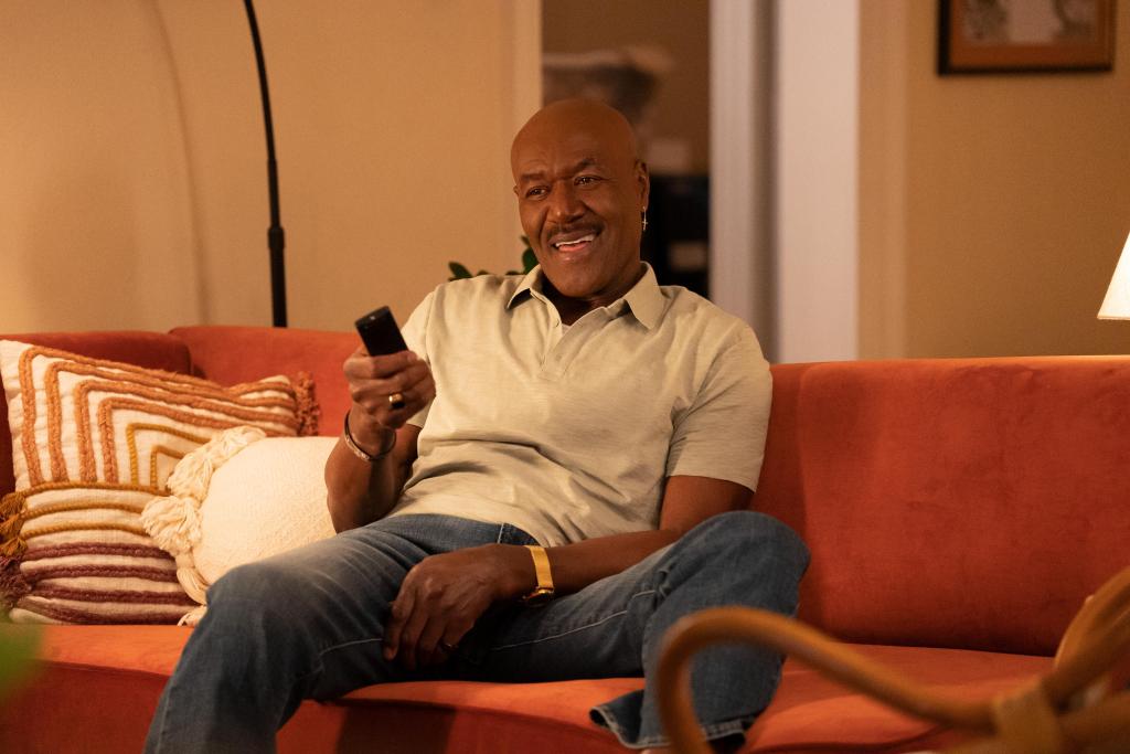 Delroy Lindo sitting on a couch and holding a TV remote, smiling. 