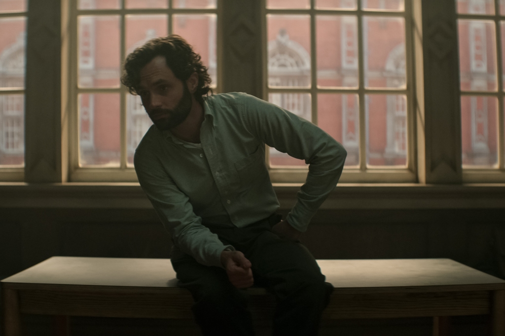 Penn Badgley sitting on a bench in a room. 