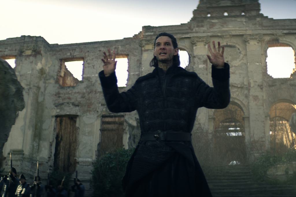 Ben Barnes with a wig, standing in a ruined building with his hands up. 