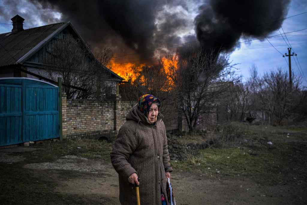 An elderly woman stands near a burning building 