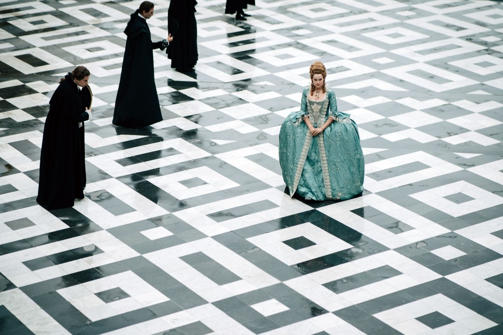 Emilia Schüle stands in the middle of a huge opulent room while people bow to her. 