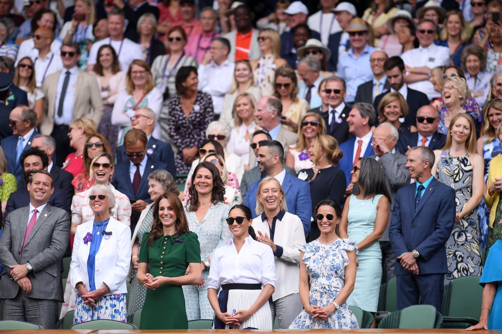 Meghan Markle with Kate Middleton and other members of the royal family at Wimbledon in 2019, standing in a crowd, smiling. 