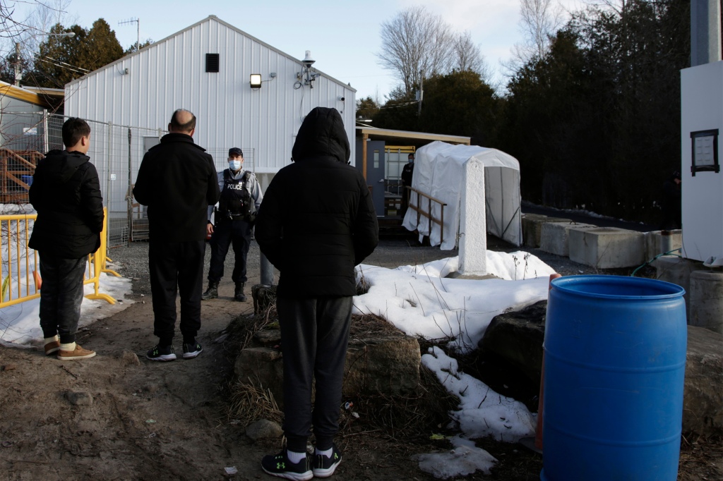 A Canadian police officer warns an Afghani migrant family that they will be arrested if they step forward into Canada at the non-official Roxham Road border crossing north of Champlain, N.Y