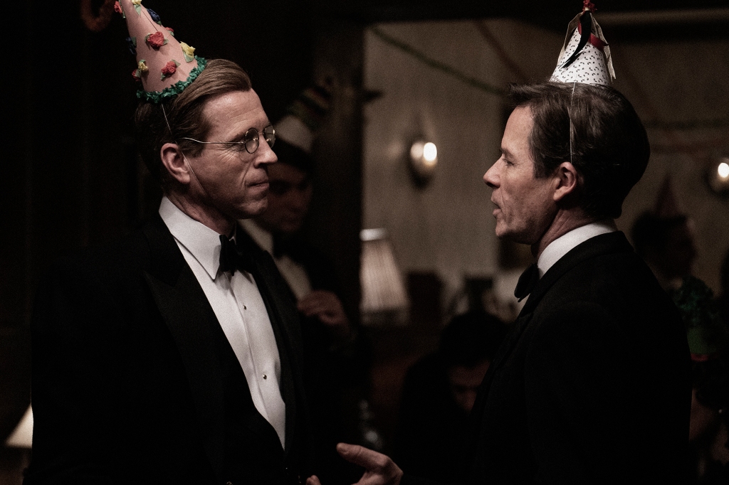 Damien Lewis and Guy Pearce as Nicholas Elliott and Kim Philby at a party in the 1940s. They're both wearing birthday hats and tuxedos and are talking to each other face-to-face.