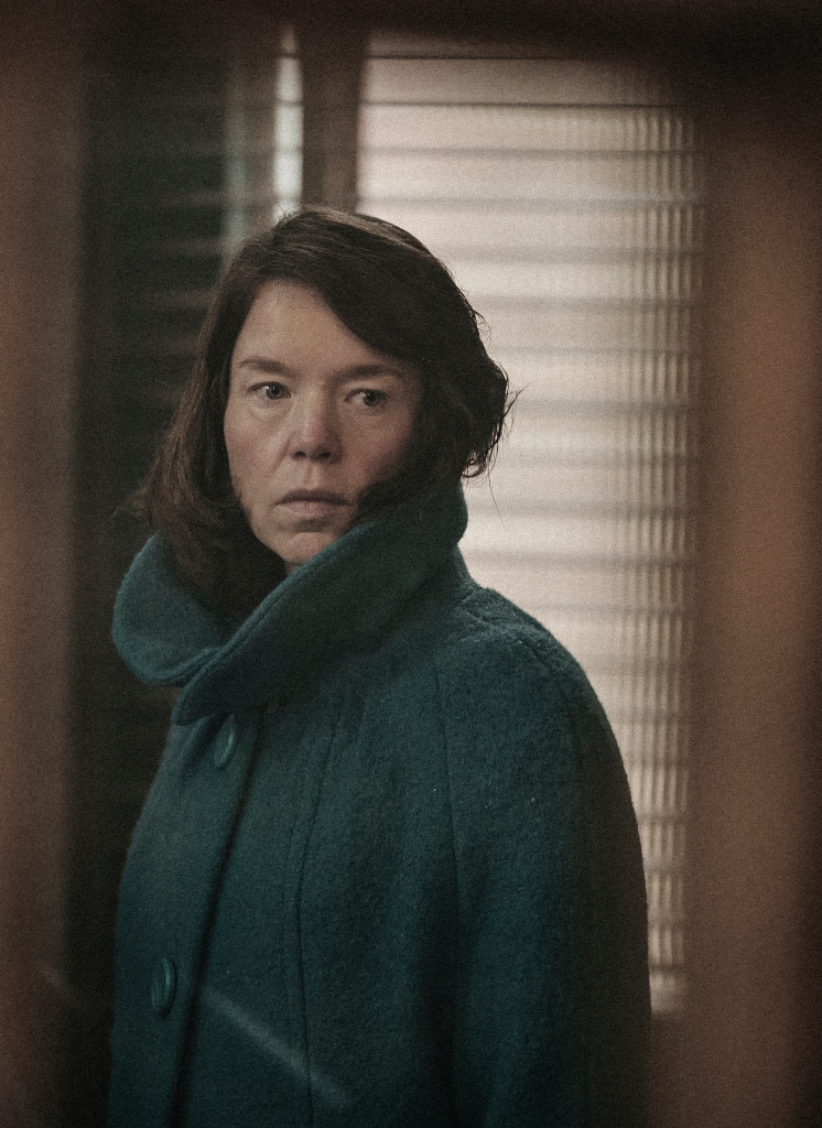 Anna Maxwell Martin as Lily Thomas. She's wearing a drab green coat and has a dour look on her face.