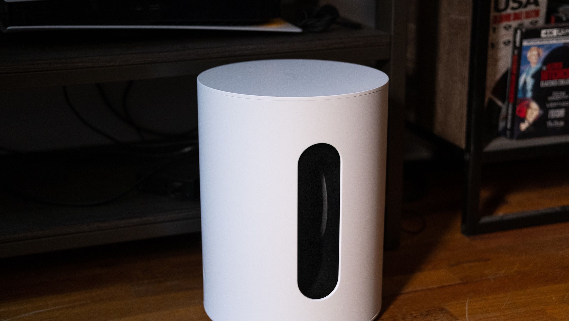 An image of the Sonos Sub Mini pictured on the floor with a TV stand and various components behind it.