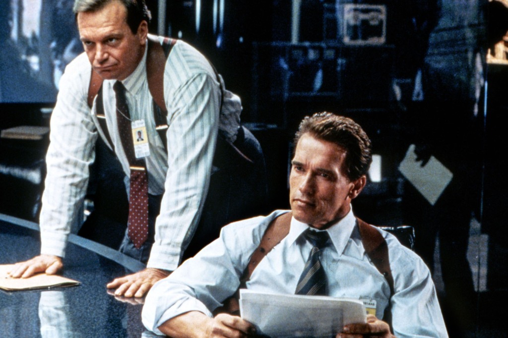 Tom Arnold and Arnold Schwarzenegger as Gib and Harry in the 1994 big-screen movie "True Lies." Gib is standing with his hands face-down on a desk while Harry is seated and staring ahead. They're both wearing gun holsters over their shoulders.