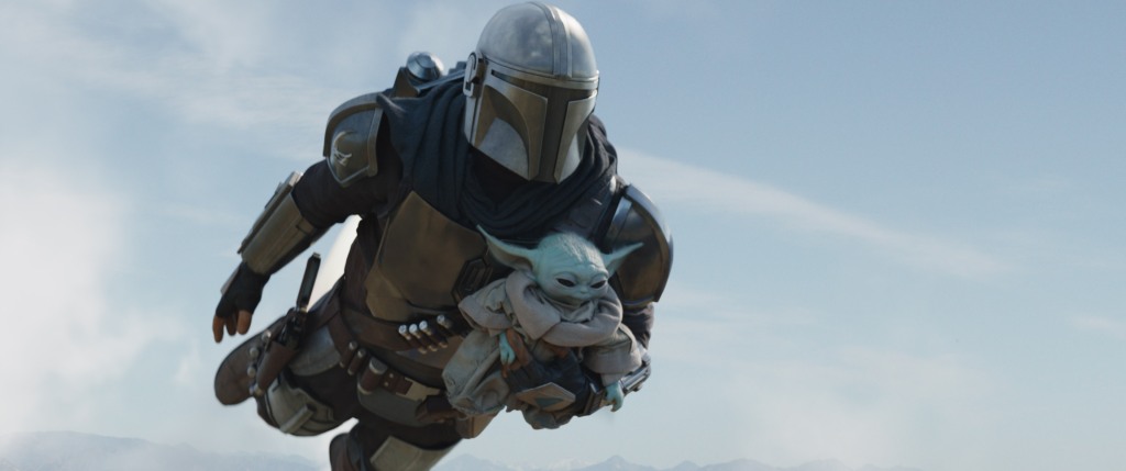 Mando in a suit and helmet, flying in the air holding Baby Yoda. 
