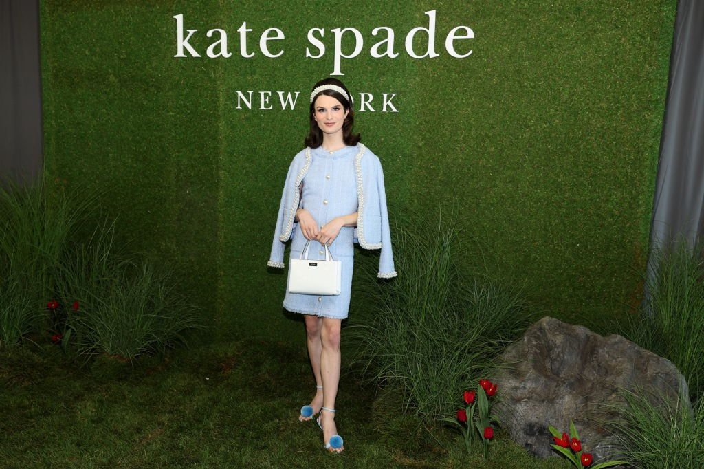 Dylan Mulvaney poses in a light blue dress and cardigan in front of a Kate Spade backdrop.