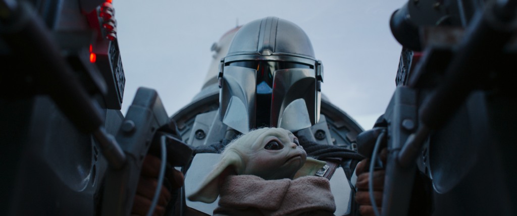 A man in a helmet and armor holding Baby Yoda. 