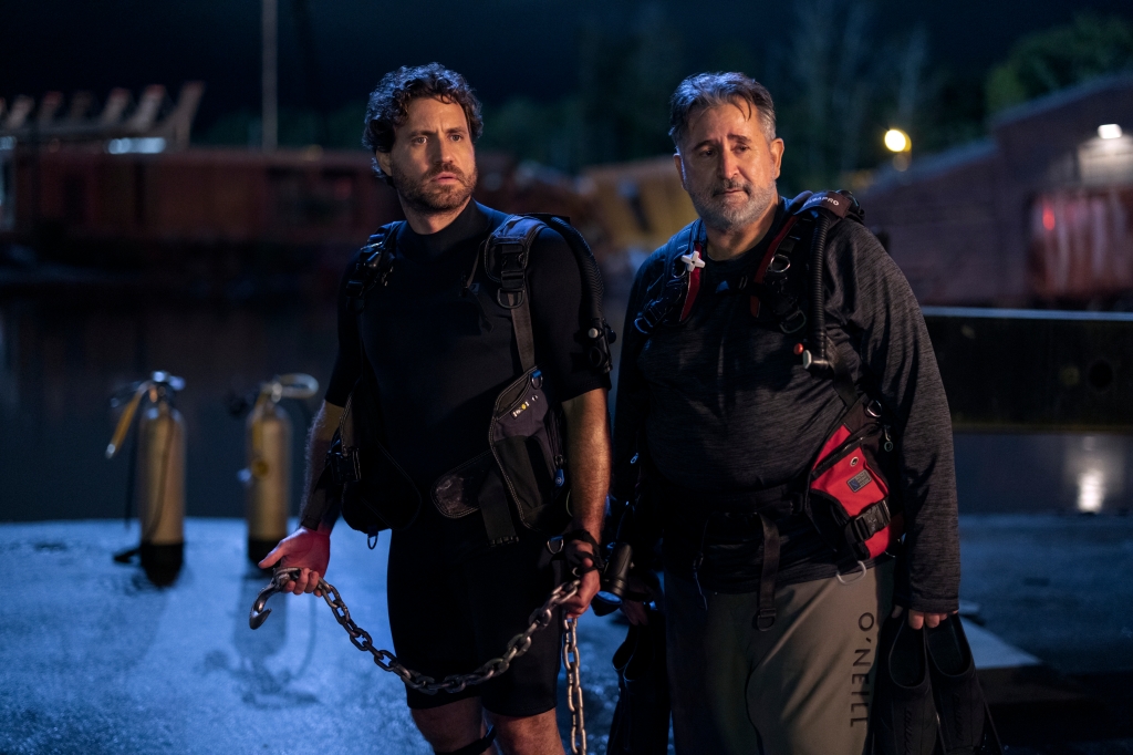 Mike (Edgar Ramirez) and his dad Sonny (Anthony LaPaglia) in "Florida Man" standing in a parking lot holding a chain wearing flak vests. 